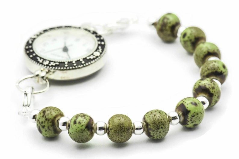 May include: A silver pocket watch with a white face and black markings attached to a green and brown beaded bracelet.