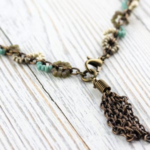 May include: A delicate necklace with a tassel made of small, linked chains. The necklace is made of a mix of small, round beads in shades of white, green, and blue. The necklace has a bronze clasp and a bronze tassel.