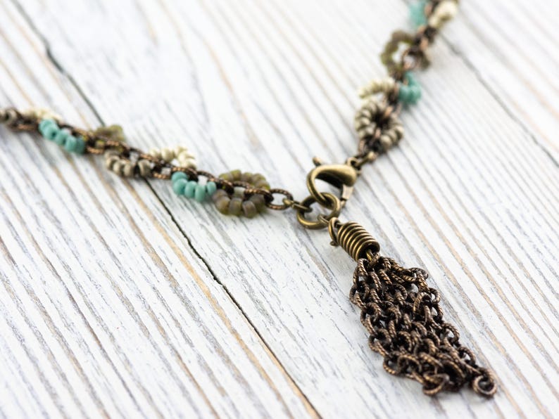 May include: A delicate necklace with a tassel made of small, linked chains. The necklace is made of a mix of small, round beads in shades of white, green, and blue. The necklace has a bronze clasp and a bronze tassel.