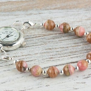 May include: A silver pocket watch with a white face and black hands is attached to a bracelet made of pink and brown striped beads and silver spacer beads.