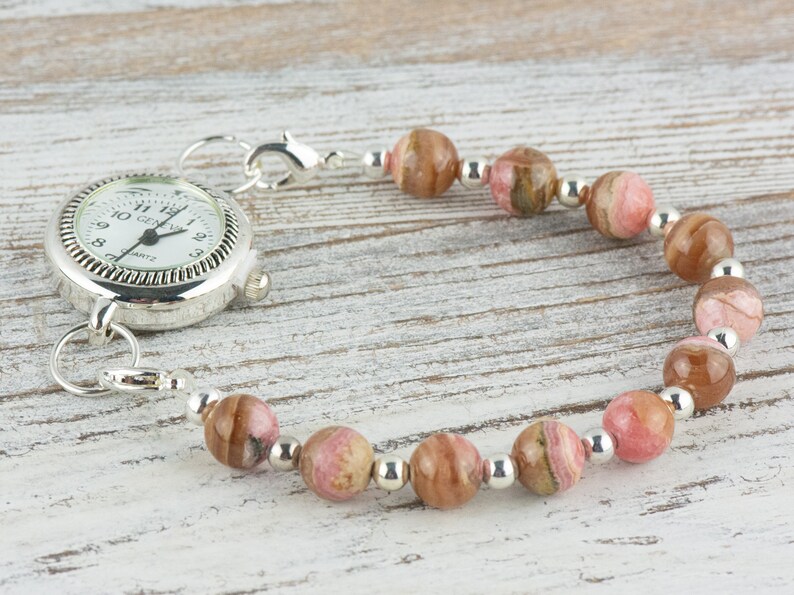 May include: A silver pocket watch with a white face and black hands is attached to a bracelet made of pink and brown striped beads and silver spacer beads.