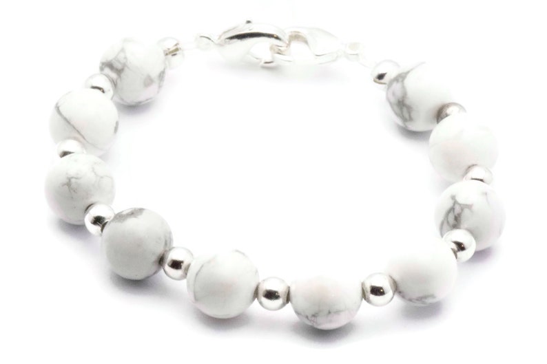 May include: A bracelet featuring white and grey marbled stone beads, interspersed with small silver-coloured beads. The bracelet is secured with a dolphin-shaped clasp. The beads are strung on a clear cord.