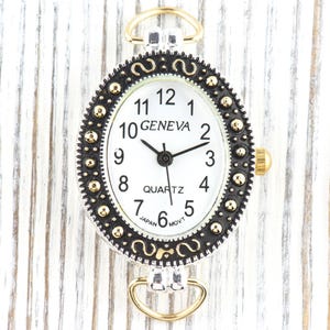May include: Oval-shaped Geneva quartz watch with a white face, black hands, and gold-tone accents. The watch features a black and gold bezel with decorative detailing and a gold crown. The watch has a silver-tone case and gold-tone loops.