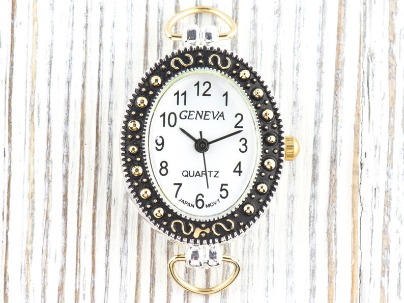 May include: Oval-shaped Geneva quartz watch with a white face, black hands, and gold-tone accents. The watch features a black and gold bezel with decorative detailing and a gold crown. The watch has a silver-tone case and gold-tone loops.