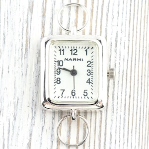 Puede incluir: Un reloj de bolsillo plateado con una esfera rectangular y una esfera blanca. La esfera del reloj muestra números y manecillas negras, con la marca "NARMI" visible. El reloj tiene dos anillos circulares para la fijación.