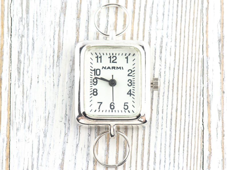 Puede incluir: Un reloj de bolsillo plateado con una esfera rectangular y una esfera blanca. La esfera del reloj muestra n&uacute;meros y manecillas negras, con la marca "NARMI" visible. El reloj tiene dos anillos circulares para la fijaci&oacute;n.