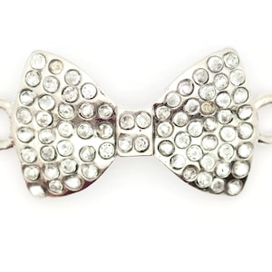 Puede incluir: Un charm de plata en forma de lazo con brillantes transparentes. El charm tiene dos lazos en los lados para sujetarlo a un collar o pulsera.