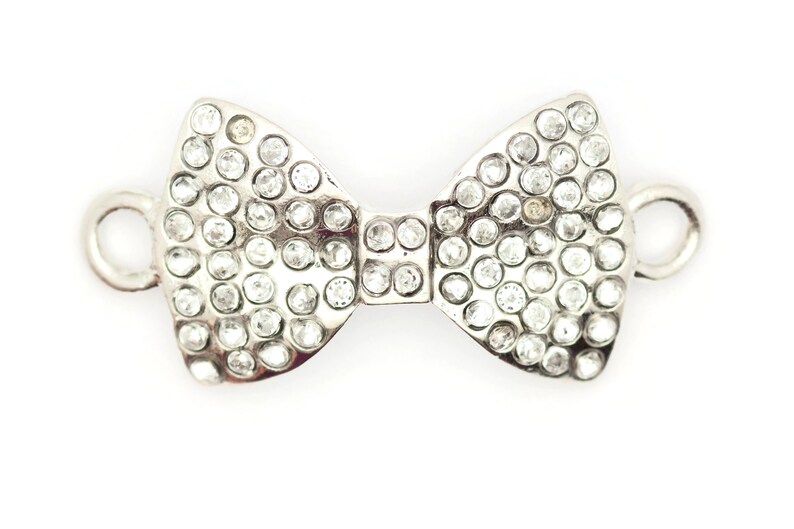 Puede incluir: Un charm de plata en forma de lazo con brillantes transparentes. El charm tiene dos lazos en los lados para sujetarlo a un collar o pulsera.