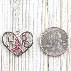 Pode incluir: Colar com pingente de cora&ccedil;&atilde;o prateado com a palavra "HOPE" e um design de fita rosa. O pingente est&aacute; ao lado de uma moeda para compara&ccedil;&atilde;o de tamanho. O colar est&aacute; em uma superf&iacute;cie de madeira branca.