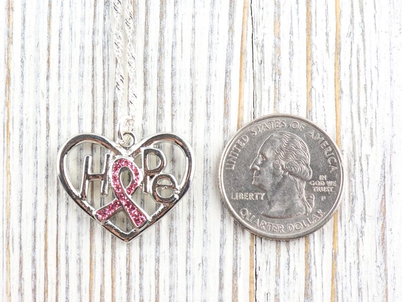 Pode incluir: Colar com pingente de cora&ccedil;&atilde;o prateado com a palavra "HOPE" e um design de fita rosa. O pingente est&aacute; ao lado de uma moeda para compara&ccedil;&atilde;o de tamanho. O colar est&aacute; em uma superf&iacute;cie de madeira branca.