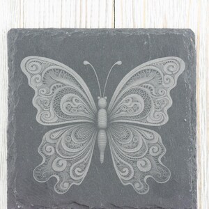 May include: A grey slate coaster featuring a white, intricate butterfly design etched into the surface.