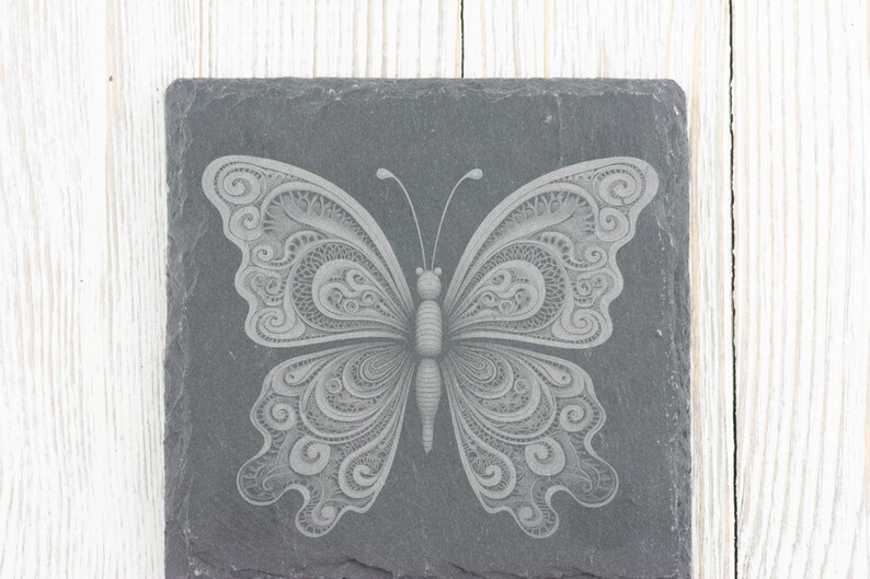 May include: A grey slate coaster featuring a white, intricate butterfly design etched into the surface.