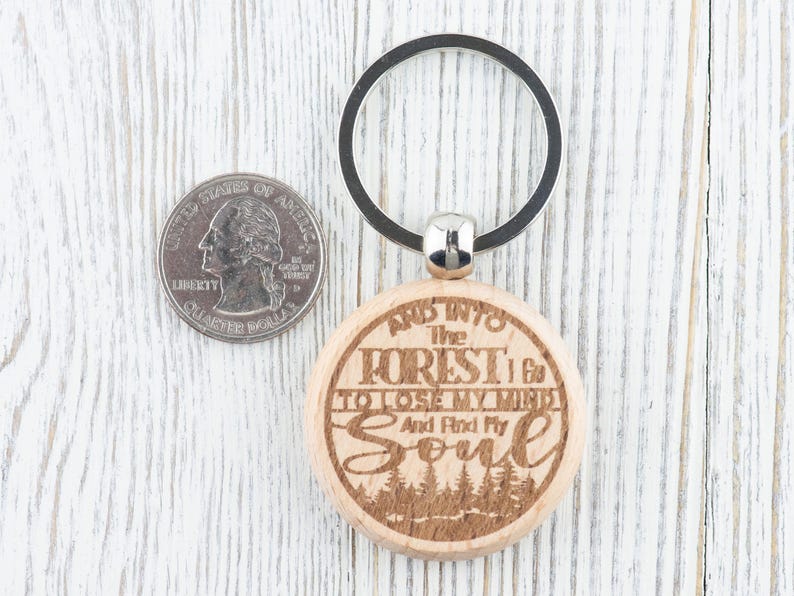 Op de afbeelding: Houten sleutelhanger met een zilveren ring. De sleutelhanger heeft een gegraveerde quote die luidt: "En de bossen in ga ik om mijn verstand te verliezen en mijn ziel te vinden".