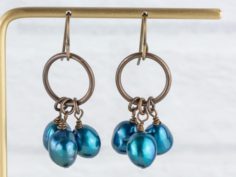 K&ouml;nnte beinhalten: Ohrringe mit bronzefarbenen Ringen und tropfenf&ouml;rmigen, teal-farbenen Perlen. Die Ohrringe h&auml;ngen an einem goldfarbenen Pr&auml;sentationsst&auml;nder. Die Perlen haben einen schimmernden, irisierenden Glanz.