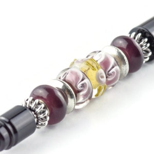 K&ouml;nnte beinhalten: Ein schwarzer Stift mit einem silbernen und violetten Perlenmuster. Die Perlen sind aus Glas und haben ein Spiralmuster.