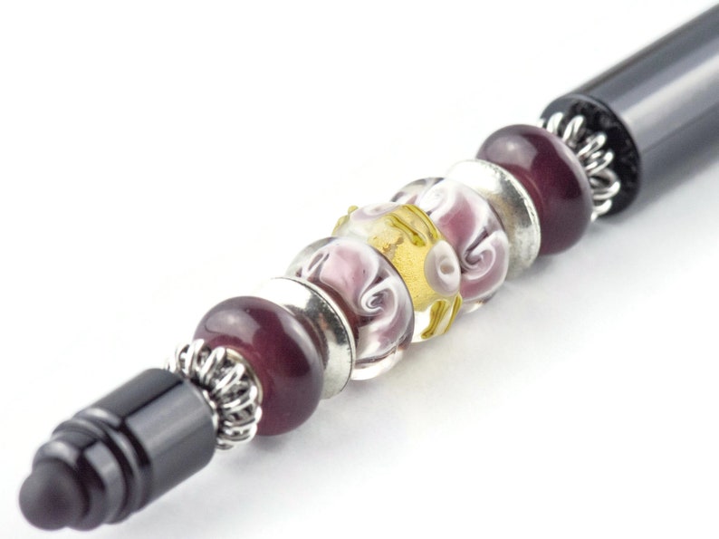 K&ouml;nnte beinhalten: Ein schwarzer Stift mit einem silbernen und violetten Perlenmuster. Die Perlen sind aus Glas und haben ein Spiralmuster.