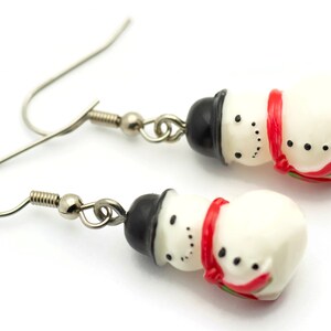 May include: A pair of silver dangle earrings with snowman charms. The snowman charms are white with black eyes and mouths, black hats, and red scarves.