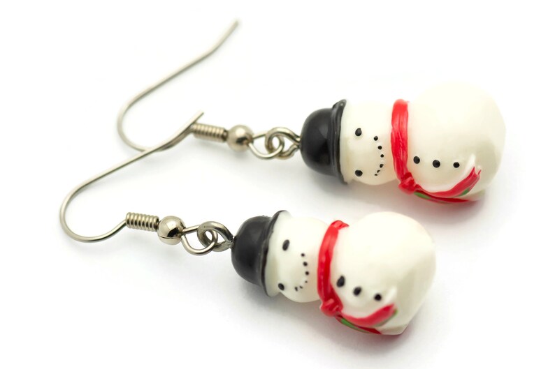 May include: A pair of silver dangle earrings with snowman charms. The snowman charms are white with black eyes and mouths, black hats, and red scarves.