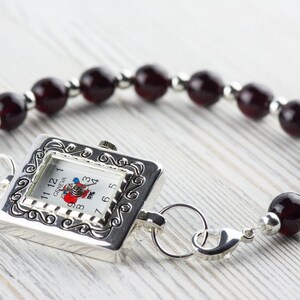 May include: A silver watch with a square face and a red and white Santa Claus design. The watch is attached to a bracelet made of dark red beads.
