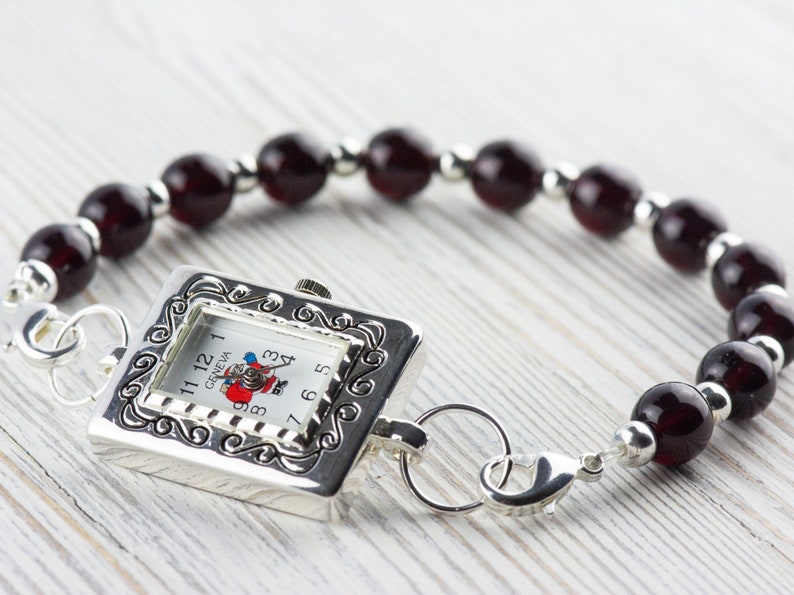 May include: A silver watch with a square face and a red and white Santa Claus design. The watch is attached to a bracelet made of dark red beads.