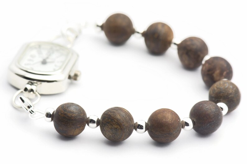 Puede incluir: Un reloj plateado con una esfera cuadrada unido a una pulsera de cuentas marrones.