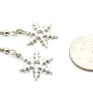 May include: A pair of silver snowflake earrings with a white enamel finish. The earrings are shown next to a US quarter for size comparison.