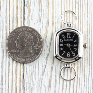 May include: A silver-toned Geneva watch with a black face and white numerals, next to a US quarter. The watch has a rectangular face with a ring at the top and bottom. The quarter is silver and shows a profile of a person.