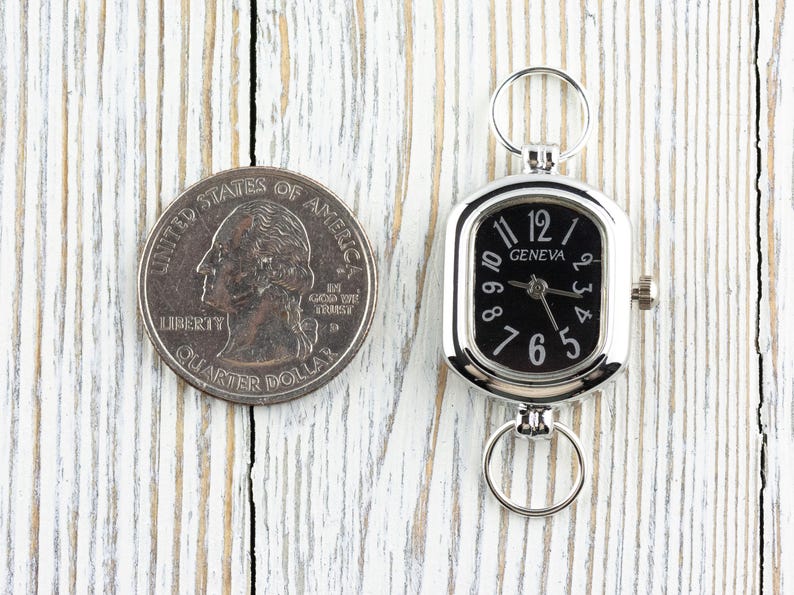 May include: A silver-toned Geneva watch with a black face and white numerals, next to a US quarter. The watch has a rectangular face with a ring at the top and bottom. The quarter is silver and shows a profile of a person.