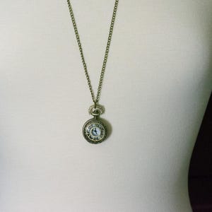 Puede incluir: Un collar con un reloj de bolsillo dorado, una esfera blanca y marcadores de hora romanos negros. El reloj tiene una cadena peque&ntilde;a y delicada.