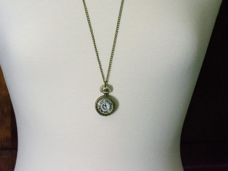 Puede incluir: Un collar con un reloj de bolsillo dorado, una esfera blanca y marcadores de hora romanos negros. El reloj tiene una cadena peque&ntilde;a y delicada.