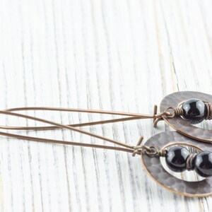 Peut inclure: Boucles d'oreilles artisanales avec des pendentifs ovales en cuivre martel&eacute;. Chaque pendentif contient deux perles noires brillantes. Les boucles d'oreilles ont de longs fils d'oreille fins en cuivre.
