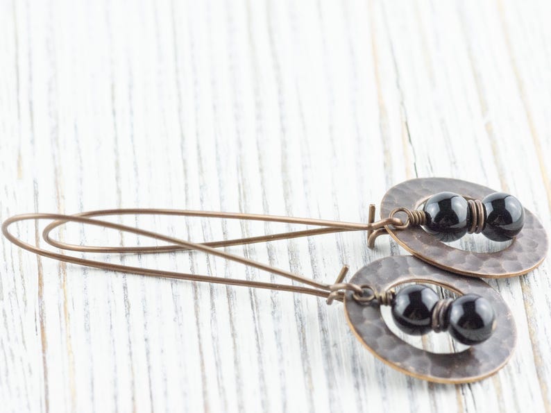 Peut inclure: Boucles d'oreilles artisanales avec des pendentifs ovales en cuivre martel&eacute;. Chaque pendentif contient deux perles noires brillantes. Les boucles d'oreilles ont de longs fils d'oreille fins en cuivre.