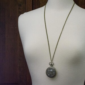 K&ouml;nnte beinhalten: Eine Halskette mit Anh&auml;nger im Vintage-Stil mit einer Taschenuhr. Die runde Uhr hat ein verziertes, detailliertes Design und h&auml;ngt an einer langen, antik-messingfarbenen Kette. Der Anh&auml;nger wird auf einer wei&szlig;en Schaufensterpuppe pr&auml;sentiert.