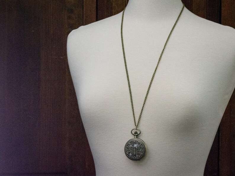 K&ouml;nnte beinhalten: Eine Halskette mit Anh&auml;nger im Vintage-Stil mit einer Taschenuhr. Die runde Uhr hat ein verziertes, detailliertes Design und h&auml;ngt an einer langen, antik-messingfarbenen Kette. Der Anh&auml;nger wird auf einer wei&szlig;en Schaufensterpuppe pr&auml;sentiert.