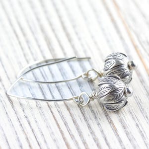 Pu&ograve; includere: Orecchini pendenti color argento con un design floreale. Gli orecchini presentano un gancio curvo e una perla strutturata dettagliata. Gli orecchini sono esposti su una superficie di legno bianco.