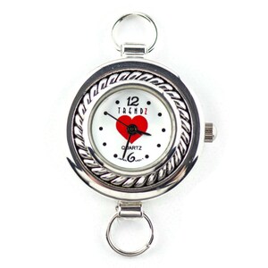 May include: Silver pocket watch with a white face, a red heart, and the word "TRENDZ". The watch has a decorative rope-like border and two ring attachments.