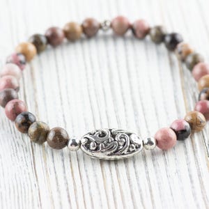 Puede incluir: Un brazalete de cuentas de color rosa y marr&oacute;n con un dije de plata con un dise&ntilde;o decorativo. El brazalete est&aacute; sobre una superficie de madera blanca.