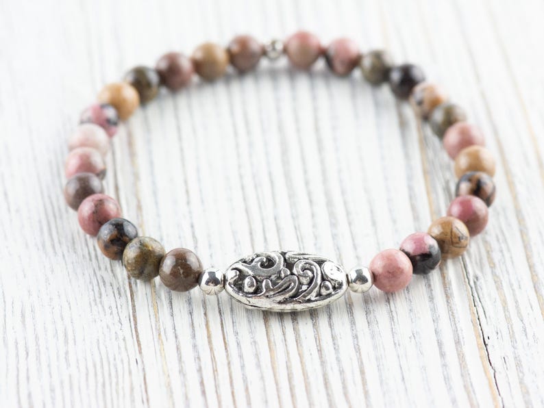 Puede incluir: Un brazalete de cuentas de color rosa y marr&oacute;n con un dije de plata con un dise&ntilde;o decorativo. El brazalete est&aacute; sobre una superficie de madera blanca.