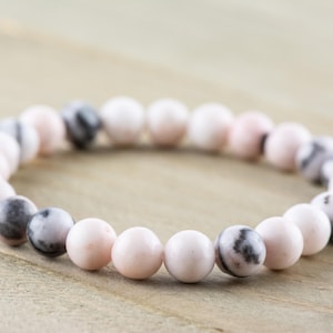 May include: A pink and white beaded bracelet with black and white marbling. The bracelet is made of round beads and is approximately 7 inches in length.