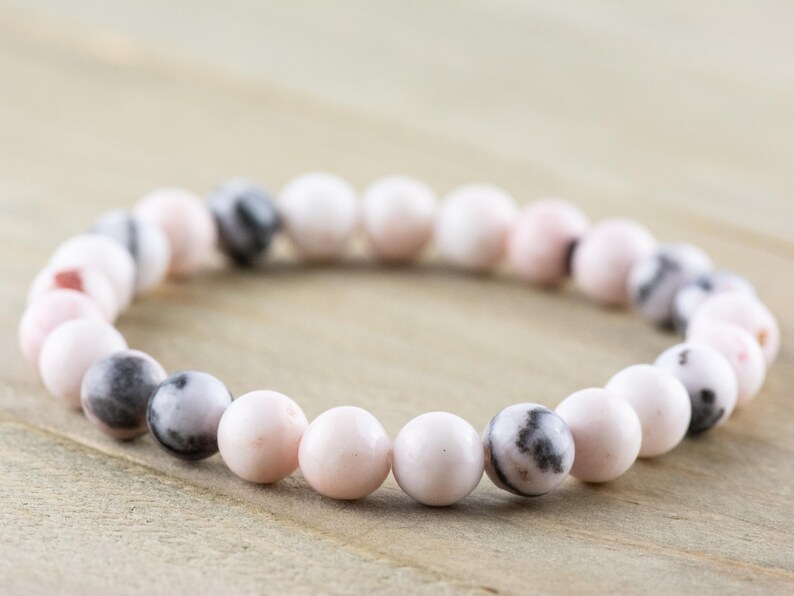 May include: A pink and white beaded bracelet with black and white marbling. The bracelet is made of round beads and is approximately 7 inches in length.