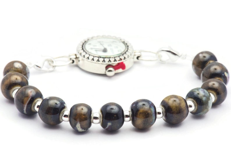 May include: A silver watch with a white face and black hands is attached to a bracelet made of brown and black ceramic beads.