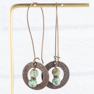 Puede incluir: Un par de pendientes largos de color cobre. Cada pendiente presenta un colgante circular martillado con un grupo de cuentas de piedra verde y marr&oacute;n. Los pendientes cuelgan de un sencillo gancho de alambre.