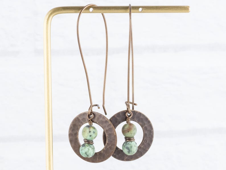 Puede incluir: Un par de pendientes largos de color cobre. Cada pendiente presenta un colgante circular martillado con un grupo de cuentas de piedra verde y marr&oacute;n. Los pendientes cuelgan de un sencillo gancho de alambre.