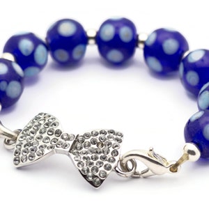 Puede incluir: Un brazalete de cuentas azules y blancas con lunares con un dije de lazo plateado y cierre.
