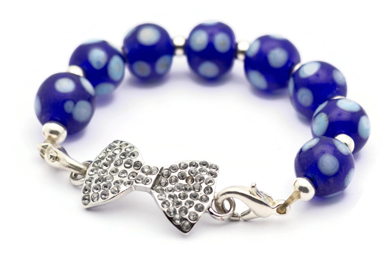 Puede incluir: Un brazalete de cuentas azules y blancas con lunares con un dije de lazo plateado y cierre.