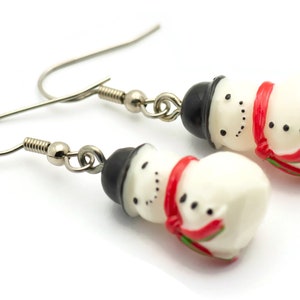 May include: A pair of silver snowman earrings with red and green accents. The snowmen are white with black hats and black eyes.
