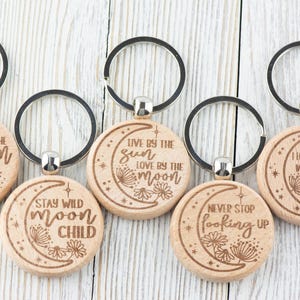 May include: Set of six wooden keychains with different moon-themed sayings. Each keychain features a crescent moon design with a different quote about the moon, such as "Shoot for the moon" and "Stay wild moon child".