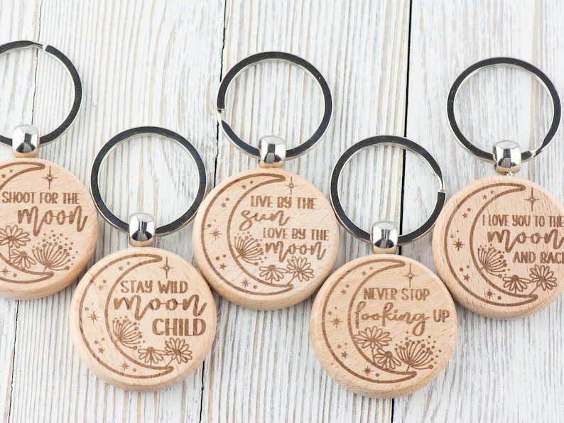 May include: Set of six wooden keychains with different moon-themed sayings. Each keychain features a crescent moon design with a different quote about the moon, such as "Shoot for the moon" and "Stay wild moon child".