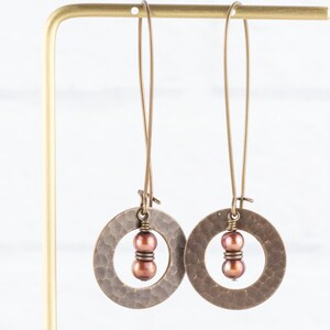 May include: A pair of bronze-colored earrings with a hammered texture. Each earring features a circular design with a cluster of two brown beads in the center. The earrings hang from long, thin wires.