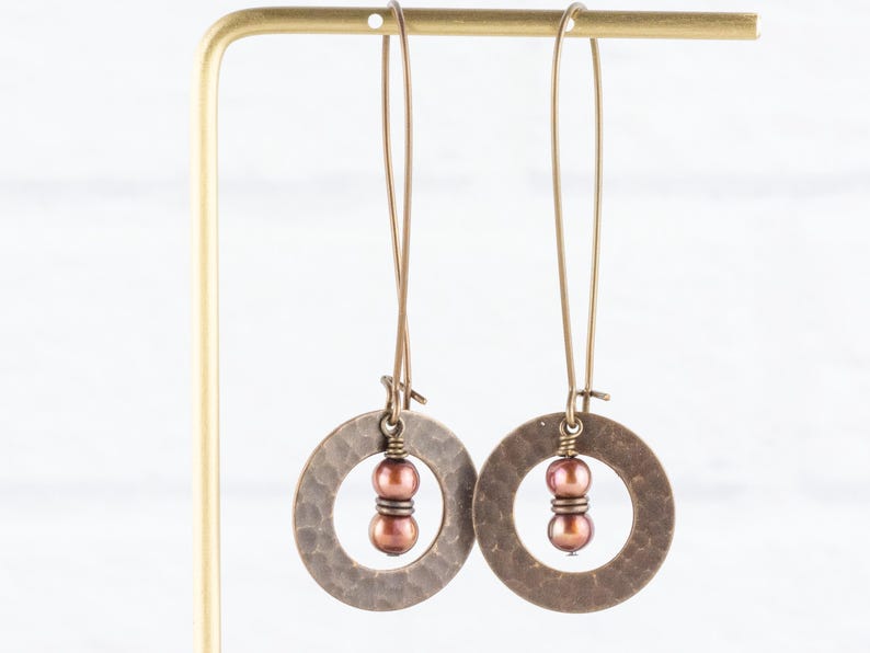May include: A pair of bronze-colored earrings with a hammered texture. Each earring features a circular design with a cluster of two brown beads in the center. The earrings hang from long, thin wires.